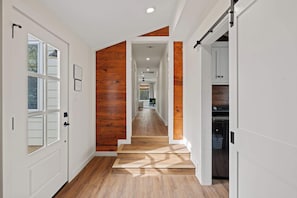 A hallway with wooden flooring and white walls, leading to a kitchen area with cabinets and appliances. - Das Morning Haus - Cozy and Spacious (Fredericksburg)