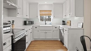 A modern kitchen with white cabinets, a stainless steel oven, a microwave, and a window with blinds.