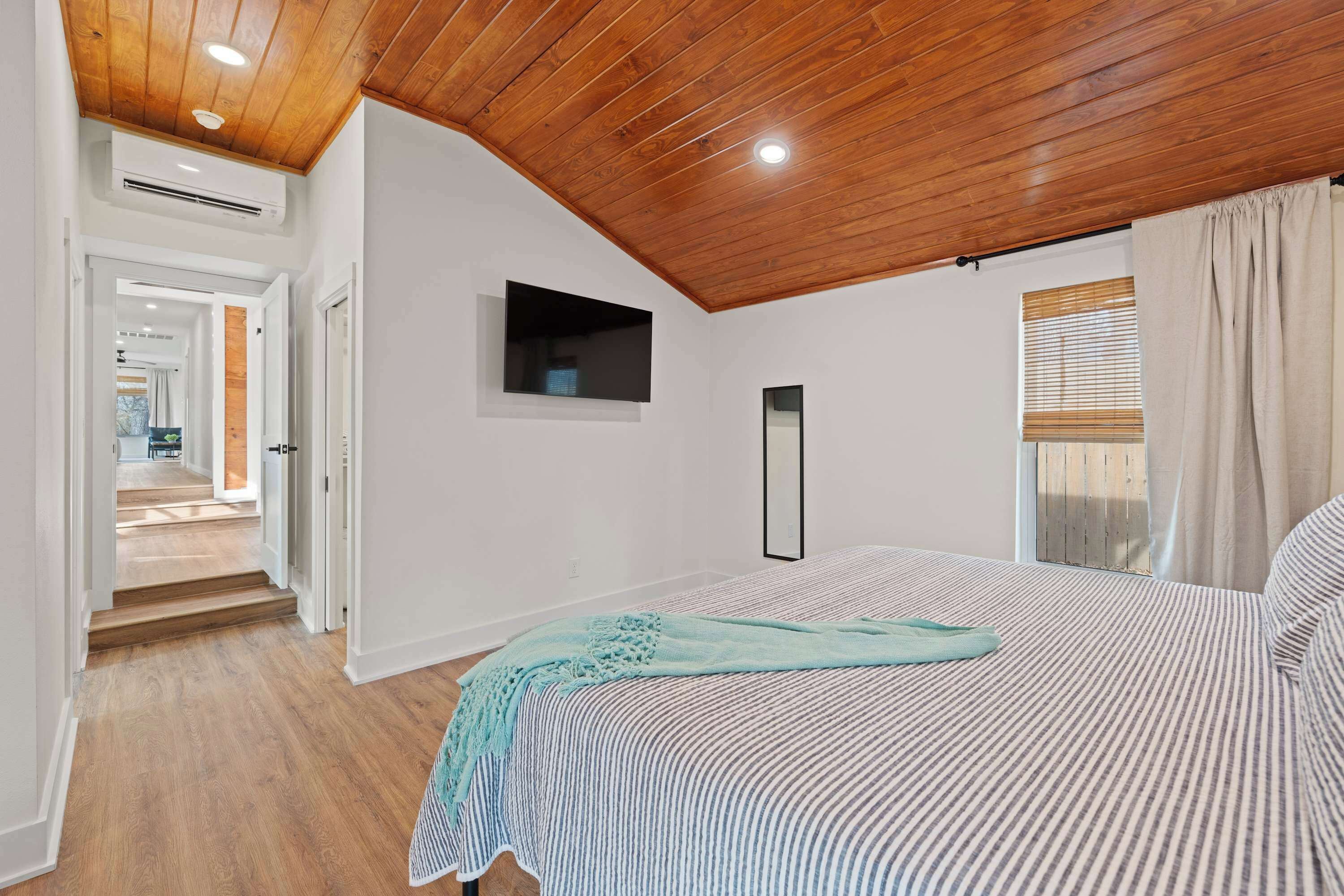 A bedroom with a wooden ceiling, a bed with striped bedding, a TV mounted on the wall, and a window with curtains.