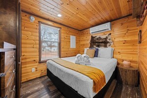 A wooden-paneled room with a bed, a window, a wall-mounted air conditioner, and a small wooden stool. - The Resort at Fredericksburg Unit 2 (Fredericksburg)