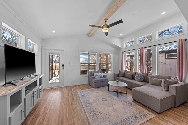 A spacious living room with a large TV, comfortable seating, a ceiling fan, and large windows offering views of the outdoors. - The Resort at Fredericksburg Unit 20 (Fredericksburg)