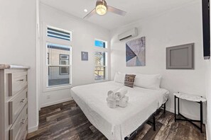 Une chambre avec un grand lit, un ventilateur de plafond, une fenêtre avec des stores, une petite table et un climatiseur fixé au mur.