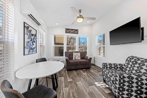 Un espace de vie compact doté d’un ventilateur de plafond, d’un téléviseur à écran plat fixé au mur, d’un canapé marron agrémenté d’un coussin à motifs floraux, d’un autre canapé à motifs, d’une table de salle à manger ronde avec des chaises, et d’une œuvre d’art représentant un papillon, encadrée et accrochée au mur.