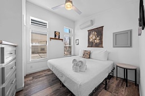 A bedroom with a large bed, a ceiling fan, a window with blinds, a wall-mounted air conditioner, and a small table. - The Resort at Fredericksburg Unit 25 (Fredericksburg)