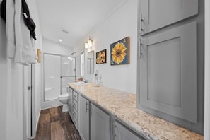 A modern bathroom with a white sink, a toilet, and a shower. There are two wall-mounted lights and a large sunflower painting. - The Resort at Fredericksburg Unit 25 (Fredericksburg)