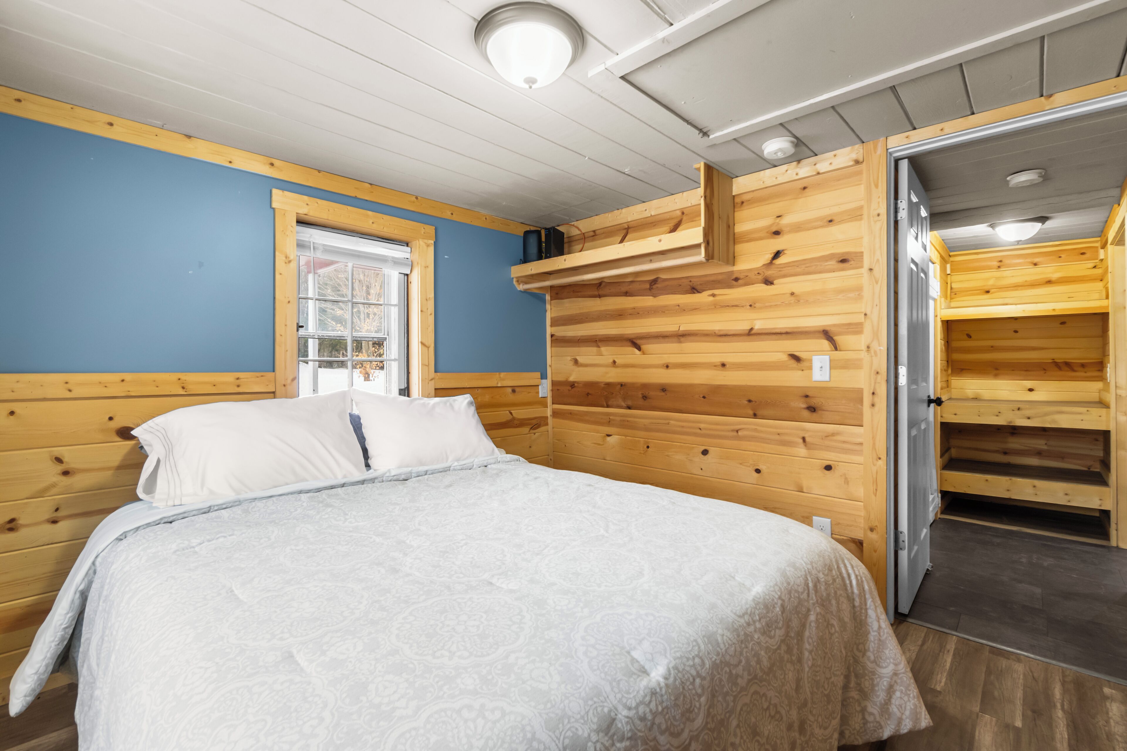 A wooden cabin room with a bed, a window, and a door leading to a loft area.