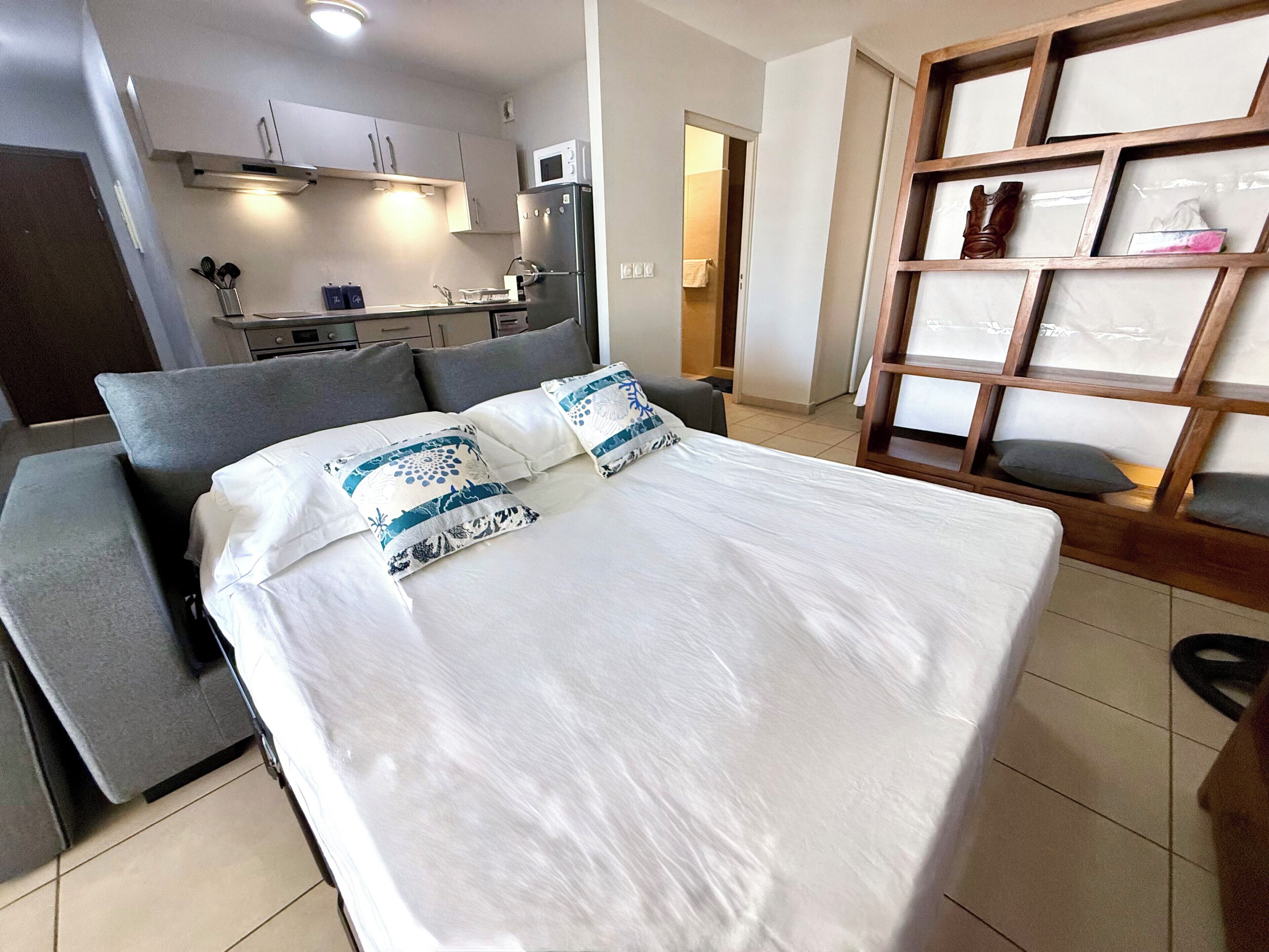 Habitación de hotel moderna con cama, sofá, zona de cocina y estante de madera con elementos decorativos.