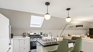 Eine kompakte Küche mit einer zentralen Kochinsel, ein Essbereich mit zwei Stühlen und ein Sofa im Wohnbereich.