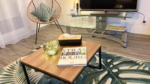 Ein modernes Wohnzimmer mit einem Glastisch, einem Korbstuhl, einem Buch mit dem Titel 'Strasbourg', einer Lampe und einem Flachbildfernseher auf einem Standfuß.