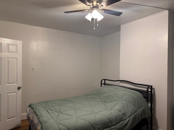 Un lit simple avec une couette verte dans une pièce aux murs blancs et dotée d’un ventilateur de plafond.