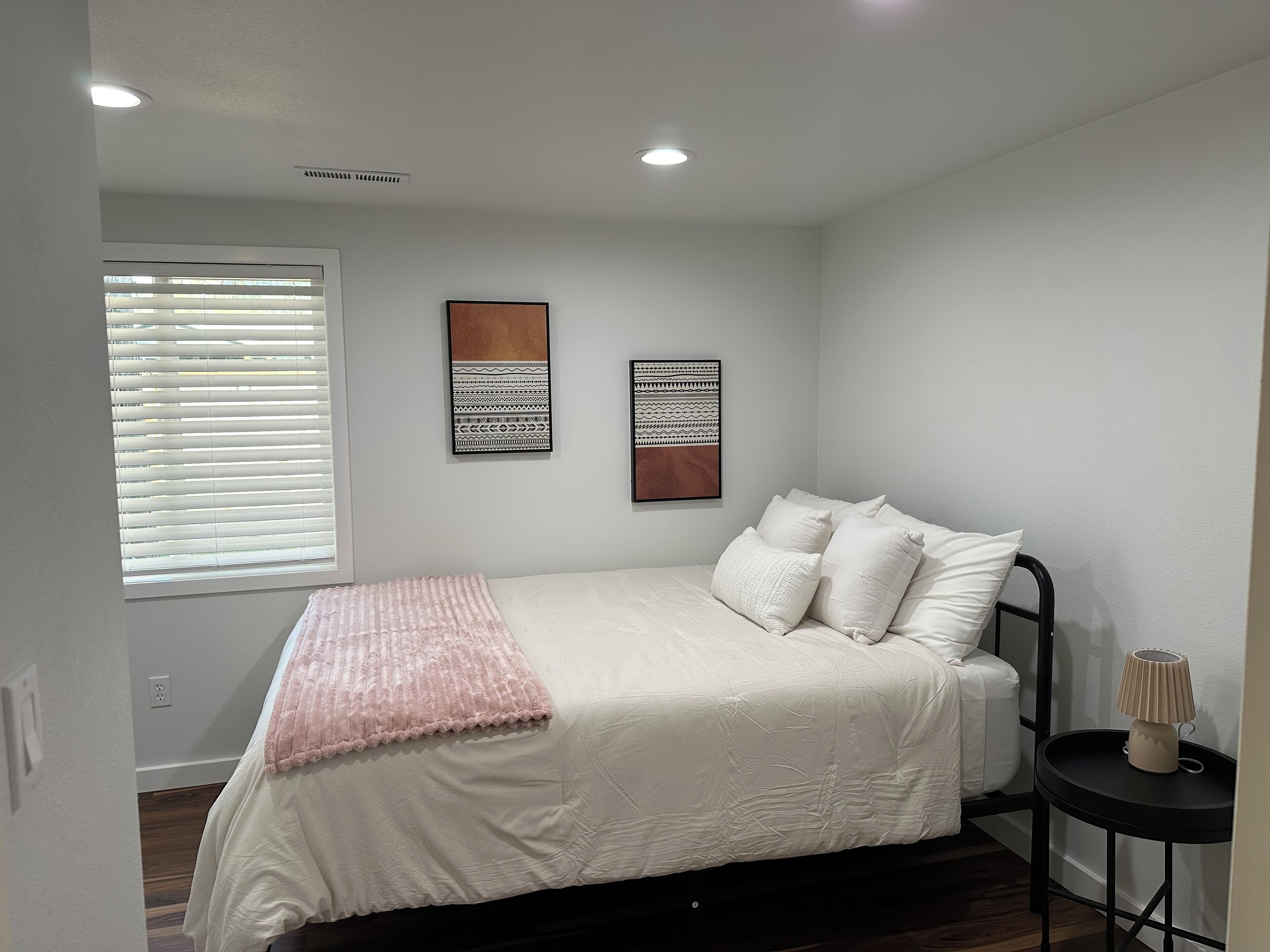 Une chambre à coucher comprenant un lit, une fenêtre avec des stores, deux œuvres d’art accrochées au mur et une table de chevet avec une lampe.