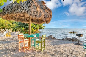 A beach setting with a thatched umbrella, a table with chairs, and a view of the sea. - Sunset, Large, Top Floor OceanFront Views, Island Condo - Key Largo (Key Largo)