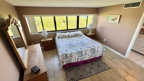 A bedroom with a large bed, a wooden dresser, a mirror, a window with a view of greenery, and a bedside table with a lamp. - Sunset, Large, Top Floor OceanFront Views, Island Condo - Key Largo (Key Largo)