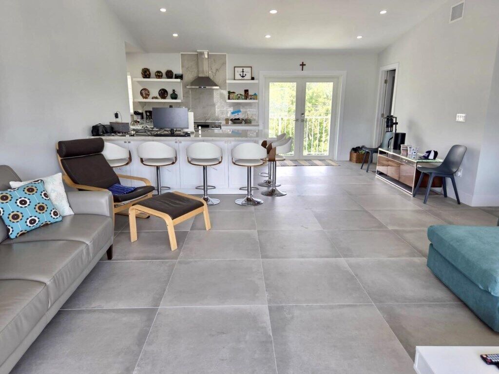 A modern living room with a grey sofa, a wooden chair, and a teal ottoman. There are three white bar stools and a kitchen area with a TV and decorative items.