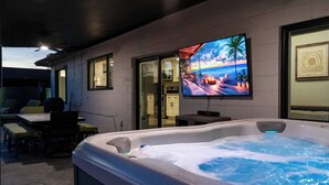 Ein Bereich mit Whirlpool, einem Fernseher, der eine Strandansicht zeigt, einer Sitzecke mit Stühlen und einer durch eine offene Tür einsehbarer Küche.