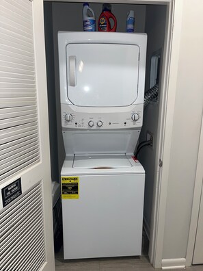 A compact washer and dryer unit with a control panel, a warning label, and detergent bottles on top. - The Comfy Stay ATL (Atlanta)