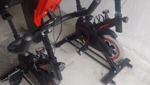 Two stationary exercise bikes with red and black components, mounted on a concrete floor with a wooden cabinet in the background.