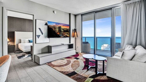 A modern hotel room with a bed, TV, sofa, and a large window with a city view. - 3BR Oceanview HYDE – Free Parking, No Resort Fees (Hollywood)