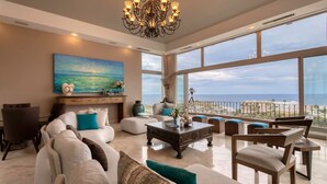 Een ruime woonkamer met een groot raam met uitzicht op een strand, een kroonluchter en een moderne zithoek.