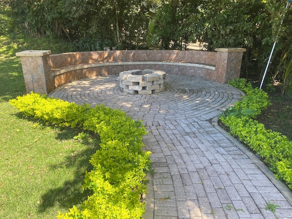 A circular brick pathway leading to a stone bench with a fire pit in the center, surrounded by lush greenery.