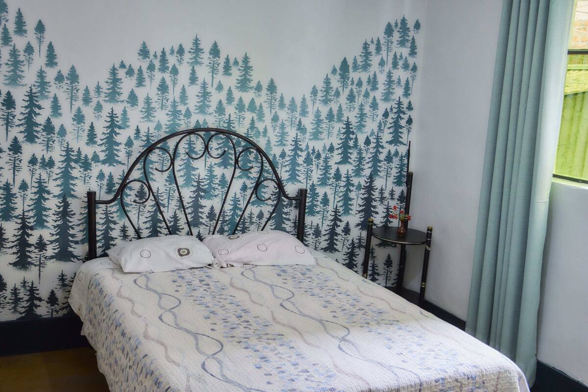 Una cama con una colcha estampada, un cabecero con papel tapiz de motivos forestales y una mesita de noche con una lámpara.