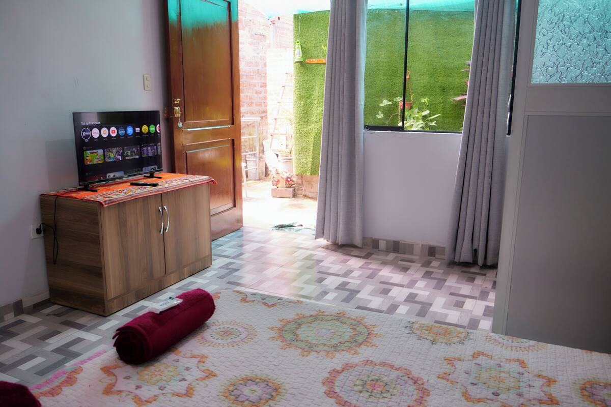 Una habitación con una televisión sobre un mueble de madera, una alfombra estampada y vistas a un césped verde a través de una ventana.