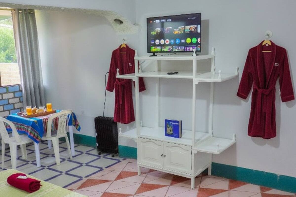 Una habitación con cama, televisor sobre un soporte, mesa con mantel y dos batas colgadas en la pared.