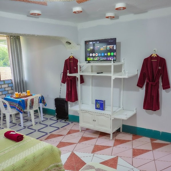 A room with a bed, a TV on a stand, a table with a tablecloth, and two robes hanging on the wall.