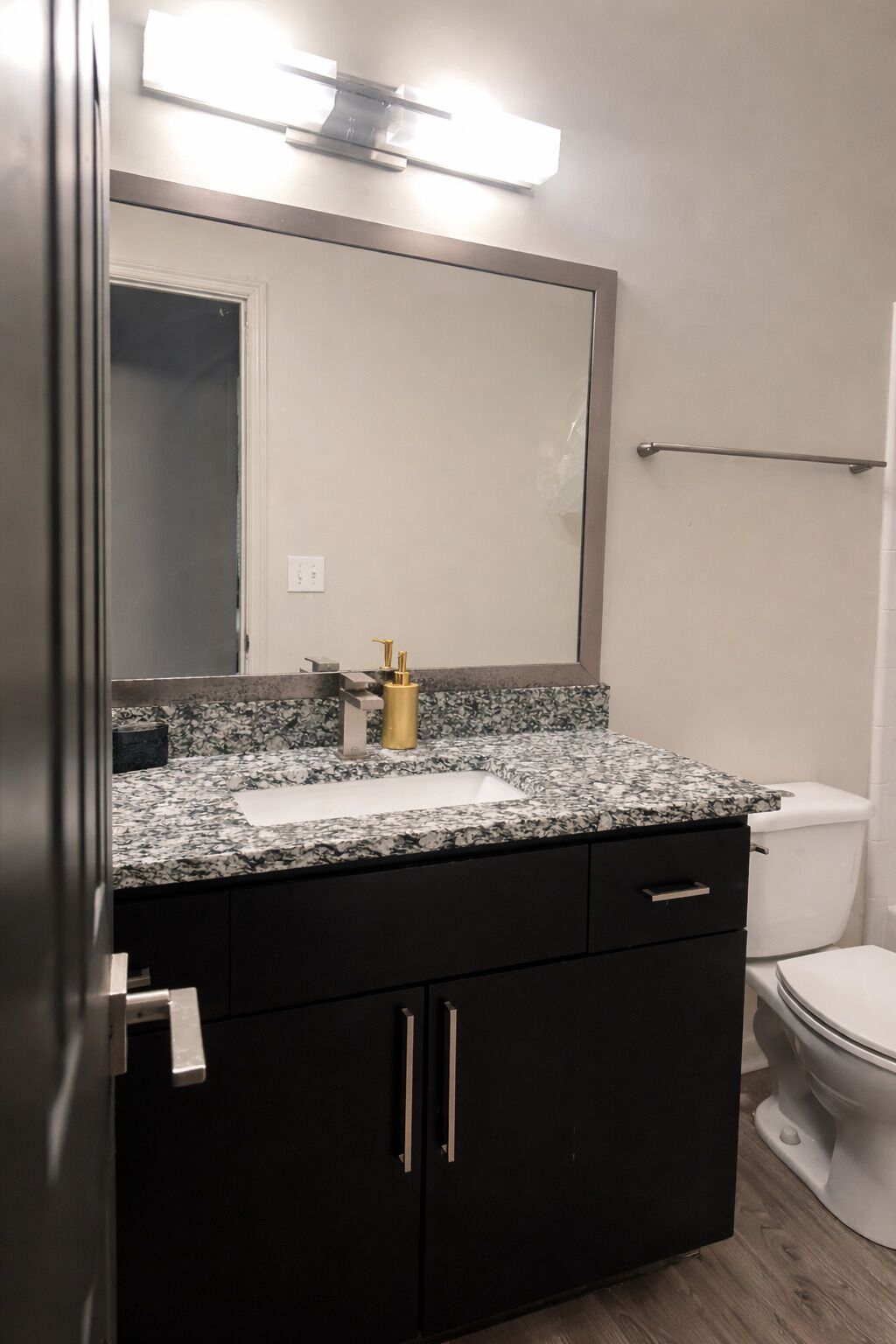A bathroom with a granite countertop, a white sink, a gold faucet, a black cabinet with silver handles, a white toilet, and a wall-mounted towel rack.