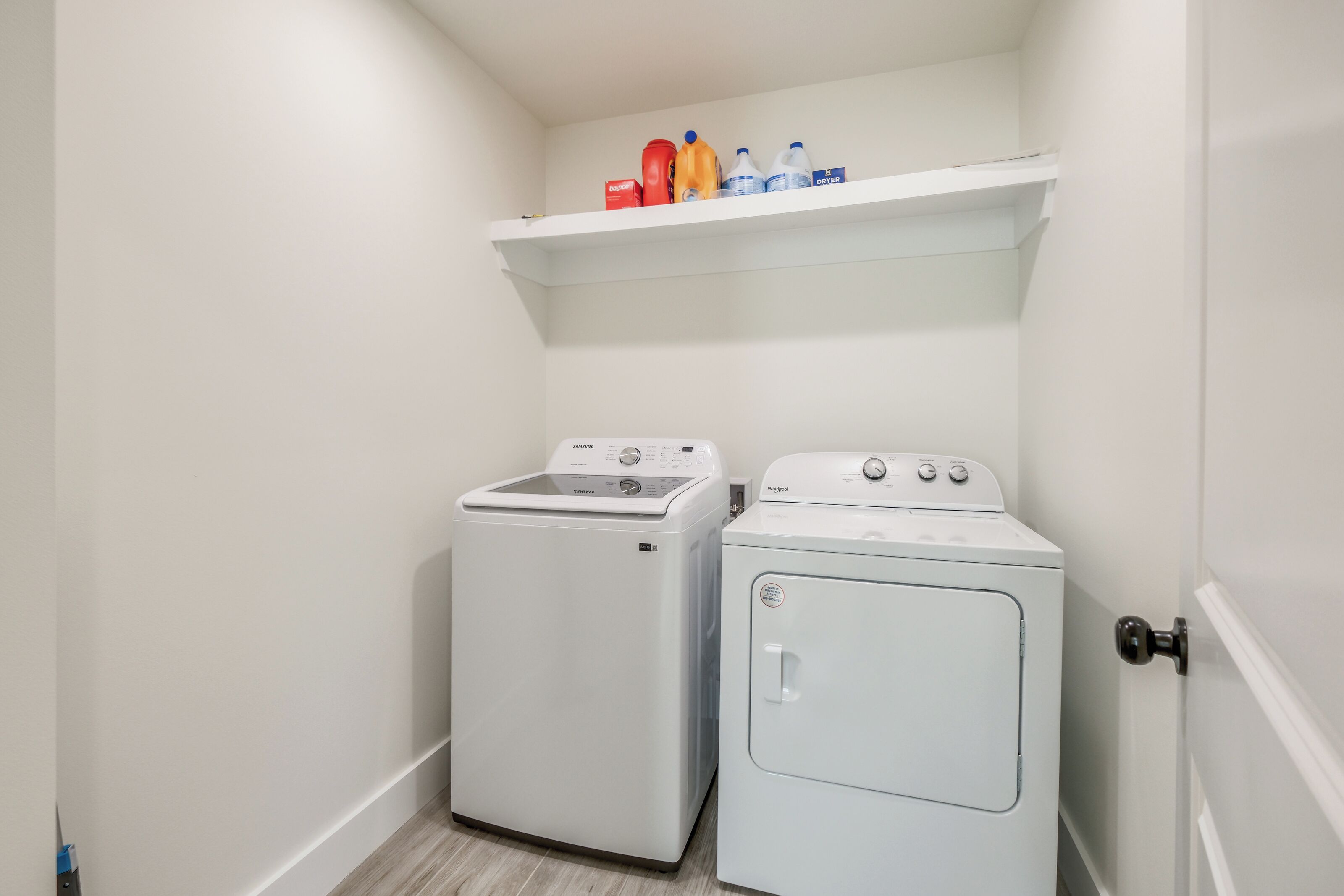 Une buanderie équipée d’un lave-linge et d’un sèche-linge, avec une étagère au-dessus où sont rangées les bouteilles de lessive, et une porte sur la droite.