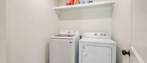 Une buanderie équipée d’un lave-linge et d’un sèche-linge, avec une étagère au-dessus où sont rangées les bouteilles de lessive, et une porte sur la droite.