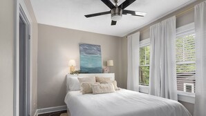 Ein Schlafzimmer mit Bett, Deckenventilator, Gemälde und Fenster mit Vorhängen.