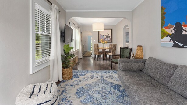 Ein modernes Wohnzimmer mit einem grauen Sofa, einem blauen Teppich mit Muster, einem Essbereich mit Tisch und Stühlen sowie einem Bild an der Wand.