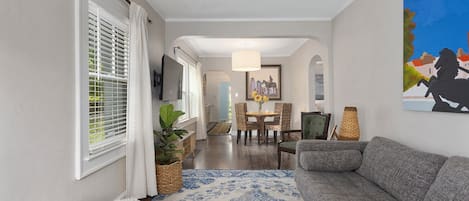 A modern living room with a grey sofa, a blue patterned rug, a dining area with a table and chairs, and a painting on the wall.