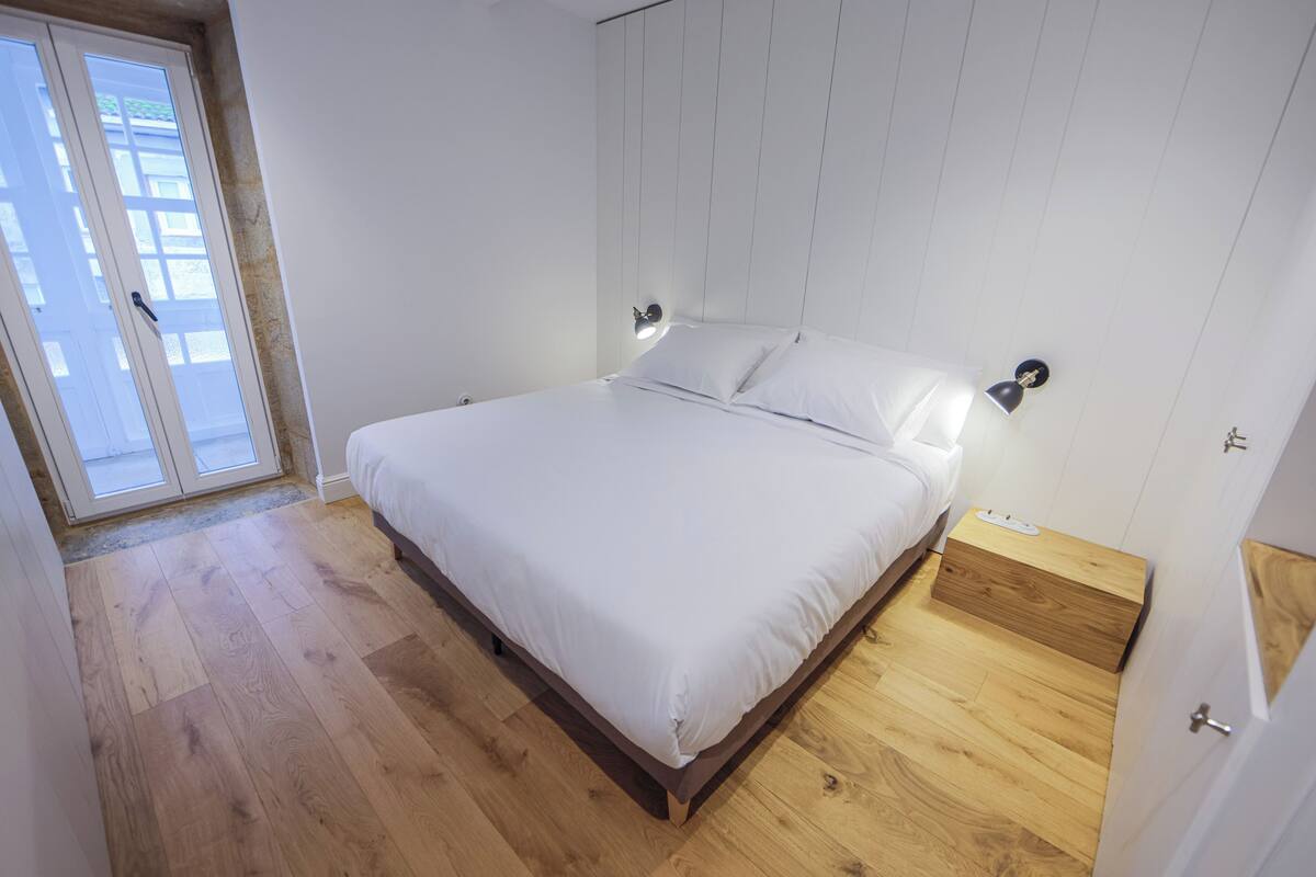 A bedroom with a wooden floor, a bed with white bedding, a nightstand, and a wall-mounted lamp.