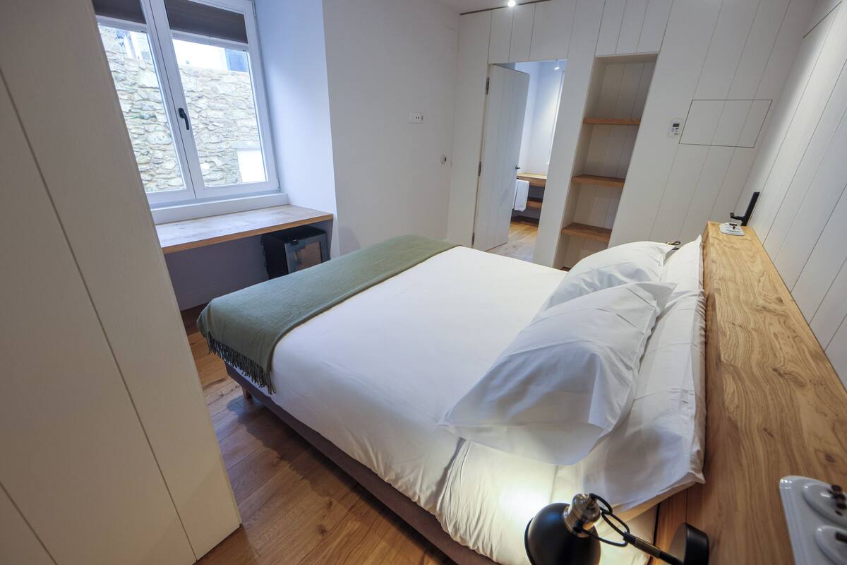 A bedroom with a bed, wooden floor, a window, a desk, and a mirror.