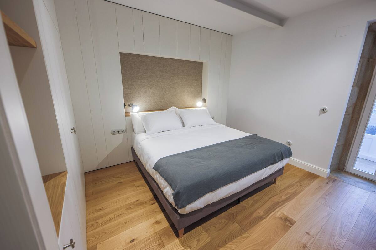 A bedroom with a bed, wooden flooring, a white wall, and a door.