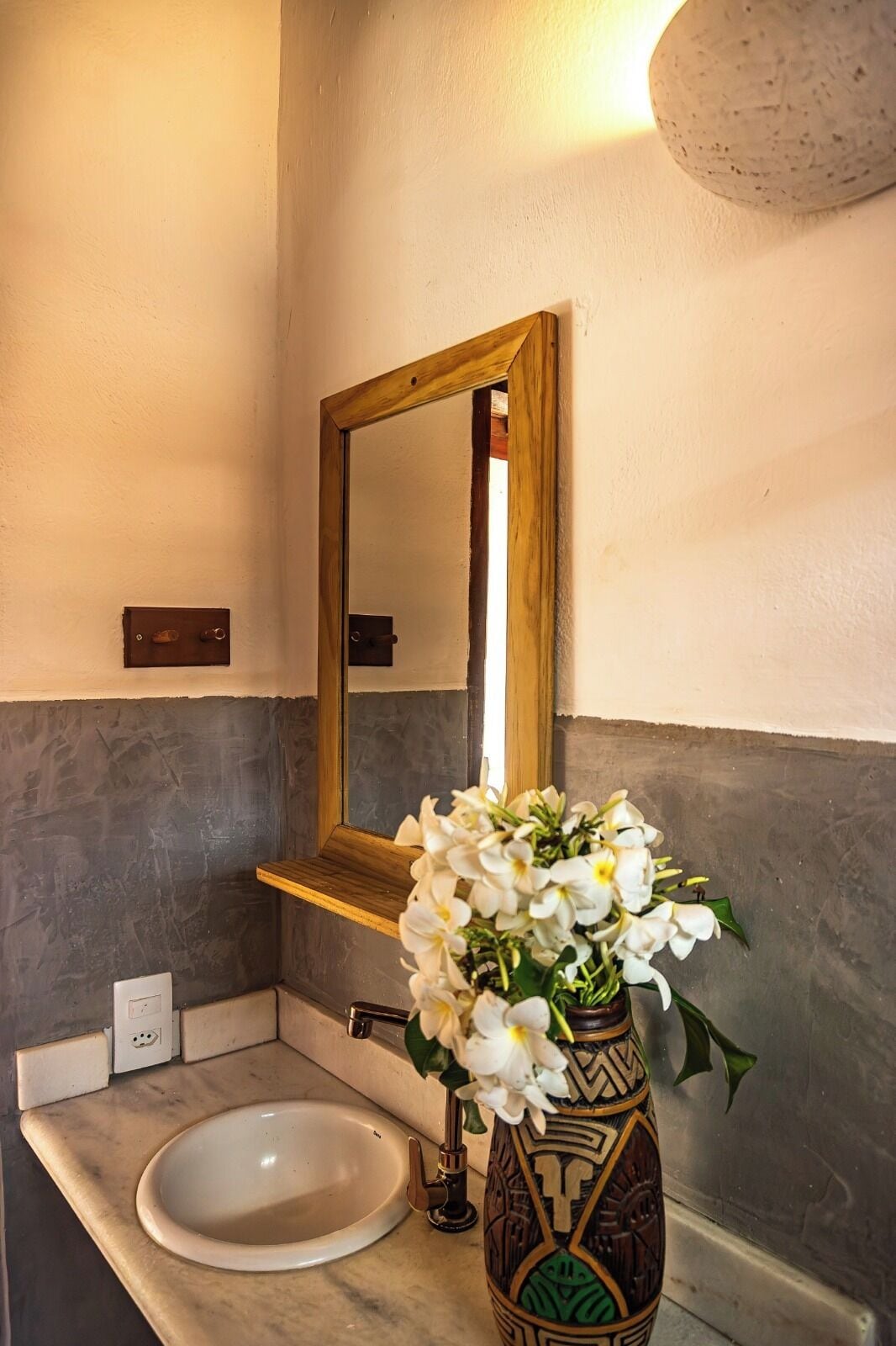 Um banheiro com pia branca, espelho com moldura de madeira e um vaso decorativo com flores brancas.