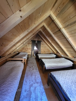 A wooden attic bedroom with four beds arranged in a row, a central pathway, and a window with curtains.