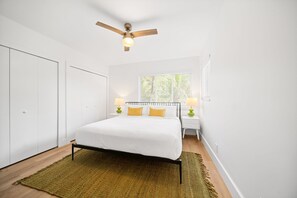 Une chambre à coucher comprenant un lit, deux tables de chevet, un ventilateur de plafond et une fenêtre avec des stores.