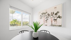 A dining area with a round table, chairs, a potted plant, a window with a view of palm trees and a building, and a framed picture on the wall. - 261 #13 Managed by Brampton Park (Deerfield Beach)