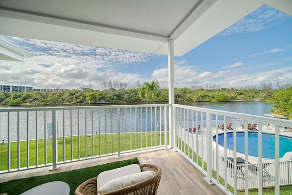 Un balcon abrité offrant une vue sur un lac, une piscine et des meubles d’extérieur.