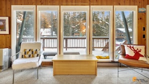 Ein gemütliches Zimmer mit Holzvertäfelung, einem weißen Stuhl, einem Holztisch und großen Fenstern mit Blick auf eine verschneite Terrasse und Bäume.