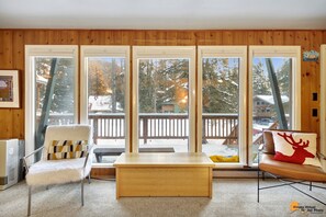 A cozy room with a wooden interior, a white chair, a wooden table, and large windows offering a view of a snowy deck and trees. - Entire cabin duplex one block from ski lifts, bike path, and hiking. (Anchorage)