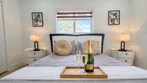 A neatly made bed with a wooden tray holding a bottle of champagne and two glasses, flanked by two bedside tables with lamps, a window with blinds, and framed floral artwork on the walls. - Stylish & Centrally Located Miami Stay / Free Parking & Fast WiFi (Miami)