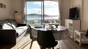 Ein modernes Wohnzimmer mit einer Couch, einem Esstisch für zwei Personen, einem Fernseher auf einem Schrank und einem Balkon mit Blick auf das Wasser.