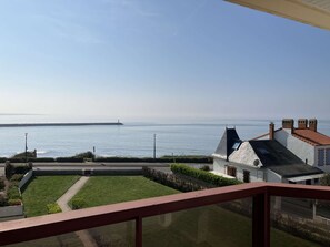 Udsigt over kysten fra en balkon med klart himmel, en fjerndrevet mole, og en hvid bygning med rødt tag.