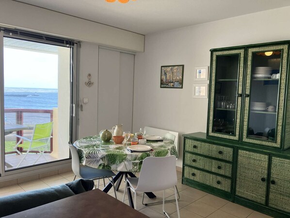 A dining area with a table set for two, a green cabinet with glass doors, and a view of the ocean through a sliding door. - Le P’tit Pilours - Saint Gilles Croix de Vie (SAINT GILLES CROIX DE VIE)