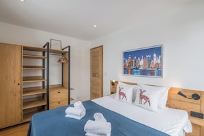 Une chambre d’hôtel avec un lit, une armoire dotée de plusieurs étagères, un tableau représentant un paysage urbain et une porte en bois.