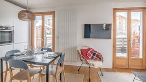 A modern kitchen with a built-in oven and microwave, a dining area with a table set for four, and a cozy seating area with a chair and a TV mounted on the wall. - Rochebrune Ski Terrace - Welkeys (Megève)